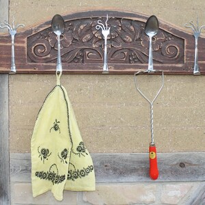 Könnte beinhalten: Ein rustikaler Holzständer mit geschnitzten Details, ausgestattet mit fünf Besteckhaken aus wiederverwendetem Besteck. Ein gelbes Geschirrtuch hängt an einem Haken, und ein Kartoffelstampfer mit rotem Griff hängt an einem anderen.