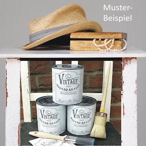 K&ouml;nnte beinhalten: Drei Dosen wei&szlig;er Vintage-Farbe mit schwarzen Etiketten und einem schwarz-wei&szlig;en Pinsel auf einem wei&szlig;en Regal. Die Dosen haben ein schwarz-wei&szlig;es Etikett mit dem Text "Vintage Paint" und einem Kranz-Design. Das Regal steht auf einem wei&szlig;en Tisch mit einer braunen Holzkiste und einem Strohhut darauf.