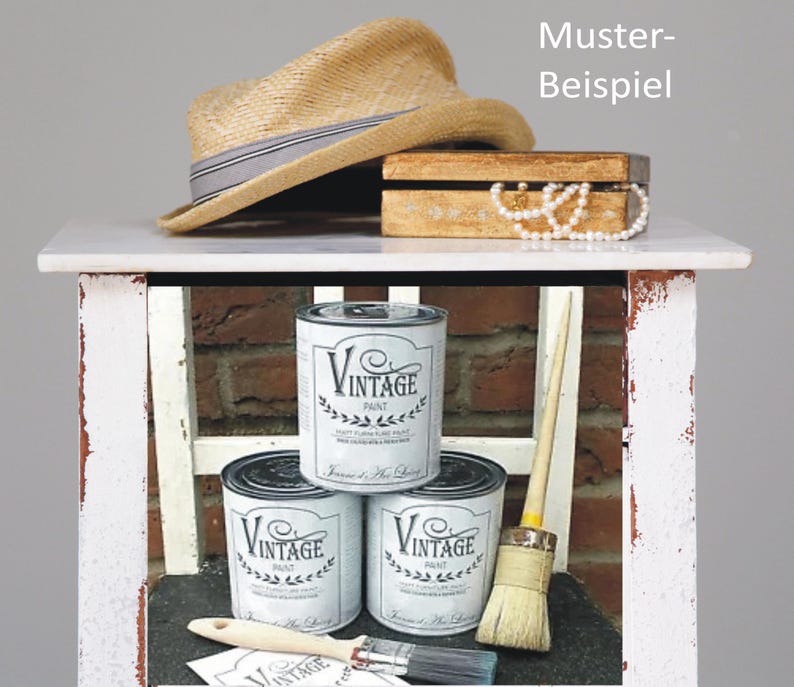 K&ouml;nnte beinhalten: Drei Dosen wei&szlig;er Vintage-Farbe mit schwarzen Etiketten und einem schwarz-wei&szlig;en Pinsel auf einem wei&szlig;en Regal. Die Dosen haben ein schwarz-wei&szlig;es Etikett mit dem Text "Vintage Paint" und einem Kranz-Design. Das Regal steht auf einem wei&szlig;en Tisch mit einer braunen Holzkiste und einem Strohhut darauf.