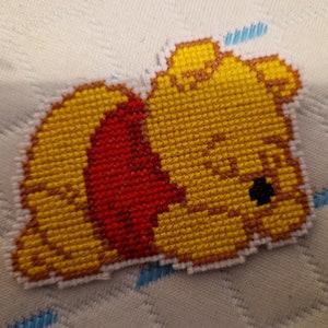 May include: A yellow and red cross-stitch of Winnie the Pooh sleeping. The cross-stitch is on a white fabric background.