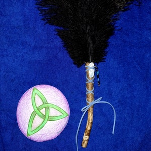 May include: A black feather smudge stick with a blue ribbon and crystal charm. The smudge stick is tied with twine and has a wooden handle. A green triquetra symbol is drawn on a pink circle.