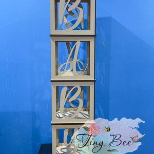 May include: Four wooden boxes stacked on top of each other, each box has a cutout of a letter spelling out the word "BABY". The boxes are a light brown colour and have a white background.