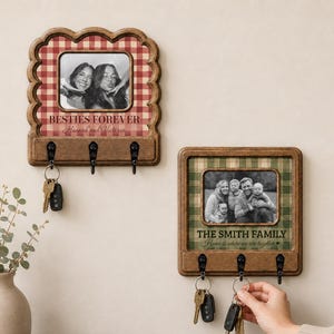 May include: Two wooden wall-mounted key holders with integrated photo frames. The top one has a red gingham background, text reading BESTIES FOREVER, and a black and white photo. The bottom one has a green plaid background, text reading THE SMITH FAMILY, and a black and white family photo. Each features three black metal hooks.