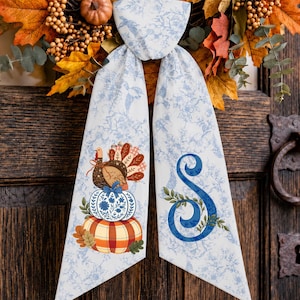May include: A decorative fabric door hanger featuring a light blue and white toile pattern. One side displays a turkey perched on stacked pumpkins, while the other side features a large blue letter S. The hanger is draped over an autumn wreath.