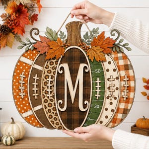 May include: A decorative wooden pumpkin wall hanging featuring a central letter M, football-themed panels with laces, a green field section with yard line markings, and various patterns including plaid, polka dots, and leopard print with autumn leaves.