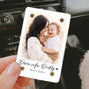 May include: A rectangular car air vent photo charm with a picture of a mother and child. The charm has the text "Drive safe, Daddy" and "Mom & Jayden" in black script. The charm has four gold-colored rivets.