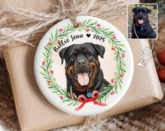 Puede incluir: Adorno navideño redondo de cerámica blanca con un retrato de un perro Rottweiler. El perro está rodeado por una corona verde con bayas rojas y un lazo rojo. El texto "Billie Jean 2025" está escrito en el adorno.