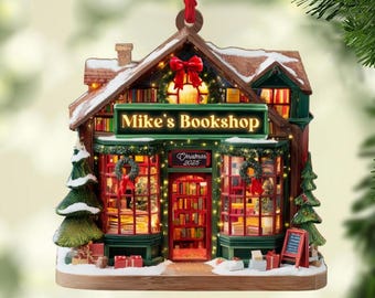 May include: A Christmas ornament shaped like "Mike's Bookshop" with a red bow, wreaths, and the text "Christmas 2025". The miniature shop has a snow-covered roof, green trim, and a red door. Presents and a small Christmas tree are at the base.