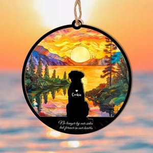 May include: A round wooden ornament with a stained glass design of a sunset over a lake. A black silhouette of a dog is in the foreground with the name "Cookie" written on its chest. The text "No longer by our sides but forever in our hearts" is written at the bottom of the ornament.