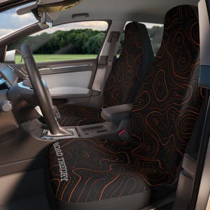 May include: A pair of car seat covers featuring a black fabric base with an orange topographic map line pattern. The text ROAD THEORY is printed in a white sans-serif font on the lower left side of the driver seat cover, visible inside a vehicle.