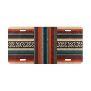 Puede incluir: Una placa de matrícula rectangular con un patrón horizontal de rayas y formas geométricas de diamante en tonos naranja, rojo, negro, crema y azul verdoso. La placa tiene cuatro ranuras de montaje, dos en la parte superior y dos en la inferior.