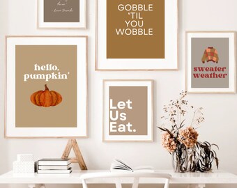 Könnte beinhalten: Fünf gerahmte Drucke mit Herbstmotiven und Texten. Die Drucke hängen an einer weißen Wand über einem weißen Schreibtisch mit einem weißen Stuhl. Die Drucke zeigen den Text "Gobble 'Til You Wobble", "Hello, Pumpkin", "Let Us Eat", "Sweater Weather" und ein Zitat über die sich verändernden Blätter.