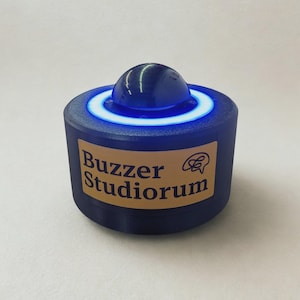 May include: A cylindrical blue buzzer device featuring a central dome button surrounded by a glowing light ring. A rectangular tan label on the front displays the text "Buzzer Studiorum" in dark blue lettering, accompanied by a small speech bubble icon.