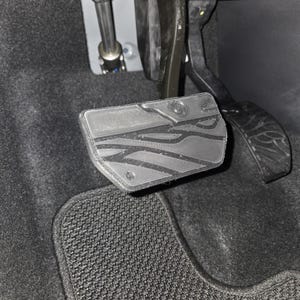 Könnte beinhalten: Nahaufnahme des Innenraums eines Autos mit einem strukturierten, grauen Gaspedal mit geometrischem Design. Das Bremspedal ist rechts sichtbar. Die Pedale sind auf einem dunkelgrauen Teppichboden angeordnet, mit einer schwarzen Fußmatte im Vordergrund.