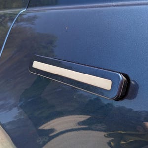 Könnte beinhalten: Nahaufnahme eines Autotürgriffs. Der Griff ist dunkelblau mit rechteckiger Form und einem hellsilbernen Akzent in der Mitte. Die Autotür ist ebenfalls dunkelblau.