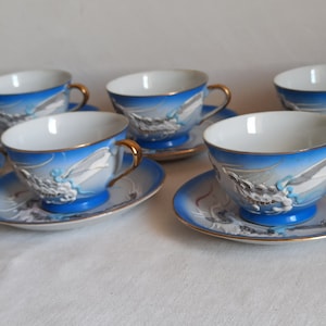 May include: Set of porcelain teacups and saucers. Each cup and saucer features a blue and white design with a dragon motif. The cups have gold handles and rims. The saucers have a matching dragon design and gold trim.