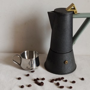 May include: A black and gray stovetop espresso maker with a light green handle and a gold-colored accent. A small stainless steel cup sits next to the coffee maker, with scattered coffee beans on the white surface.