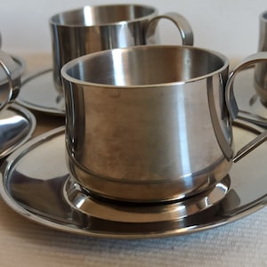 May include: Set of stainless steel coffee cups and saucers. The cups have a rounded shape with a curved handle. The saucers are oval-shaped. The set is made of shiny, polished metal, reflecting the light. The cups and saucers are arranged on a white surface.