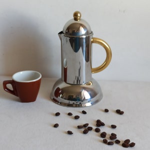 Op de afbeelding: Een roestvrijstalen espressomaker met een gouden handvat en bovenkant, naast een kleine bruine espressokop met een witte binnenkant. Verspreide koffiebonen completeren de scène, wat wijst op een product met koffiethema.