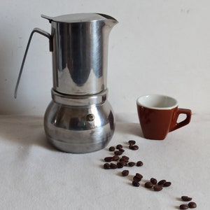 May include: A stainless steel stovetop espresso maker with a small brown coffee cup and scattered coffee beans. The espresso maker has a round base and a top with a handle. The cup is brown with a white interior.