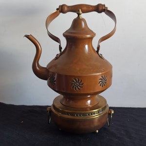 May include: A copper-colored vintage kettle with a wooden handle and a decorative floral pattern. The kettle has a spout, a lid, and a brass base. The handle is made of wood and the base has small feet.