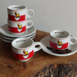 May include: A set of espresso cups and saucers. The cups are white with a red band and a coffee cup graphic. The word "SAICAR" is printed on the cups. The set includes coffee beans on a saucer, all displayed on a wooden surface.
