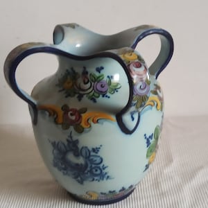 May include: A light blue ceramic pitcher with two handles and a wide spout. The pitcher is decorated with hand-painted floral designs in shades of blue, purple, yellow, and green. The handles and rim are outlined in dark blue.