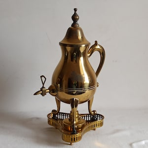 May include: A vintage brass coffee urn with a spigot, handle, and lid. The urn sits on a decorative base with three legs. The brass has a polished finish, reflecting the light. The overall design is ornate and classic.