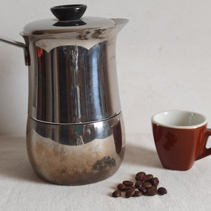 Puede incluir: Una cafetera italiana de acero inoxidable con asa y tapa negras, junto a una pequeña taza de espresso marrón con interior blanco. También hay un pequeño montón de granos de café. Los elementos están sobre una superficie blanca.