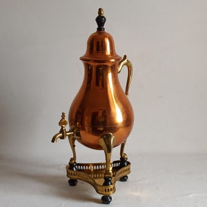 May include: A copper-colored antique coffee urn with brass accents. The urn has a bulbous body, a spigot, and a lid with a black finial. It sits on a brass stand with decorative feet.