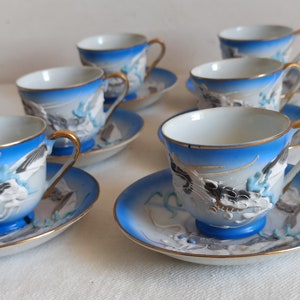 May include: Set of six porcelain teacups and saucers. Each cup and saucer features a blue and white design with gold trim. The cups have a dragon motif and gold handles. The saucers have a matching design.