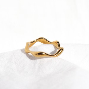 May include: A gold-colored ring with a wavy, organic design. The ring has a smooth, polished finish and appears to be made of metal. The ring is set against a white background.