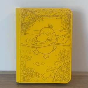 May include: A bright yellow card binder featuring a debossed line art illustration of a duck-like creature swimming in a pond with aquatic plants and rocks. The bottom left corner displays the embossed text Vault X in a clean, sans-serif font.