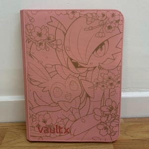 May include: Pink card binder with a gold embossed design featuring two cartoon characters and cherry blossoms. The text "vault x" is printed in gold at the bottom left corner.