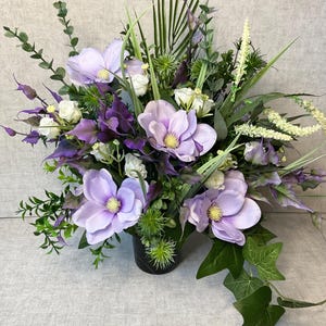 May include: A floral display with large, pale purple magnolia flowers, white roses, and diverse green foliage. The arrangement is in a black vase, with trailing ivy adding a touch of elegance. The overall effect is a sophisticated floral display.