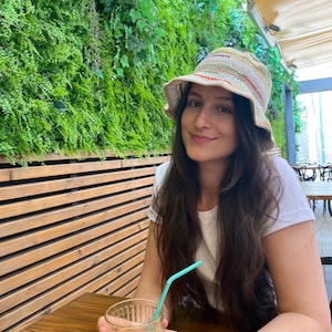 May include: A woman wearing a white shirt and a multi-colored crocheted bucket hat sits at a wooden table with a glass of orange juice and a blue straw.