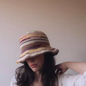 May include: A woman wearing a crocheted bucket hat with brown, orange, yellow, and white stripes. The hat is pulled down over her forehead, and she is looking down.