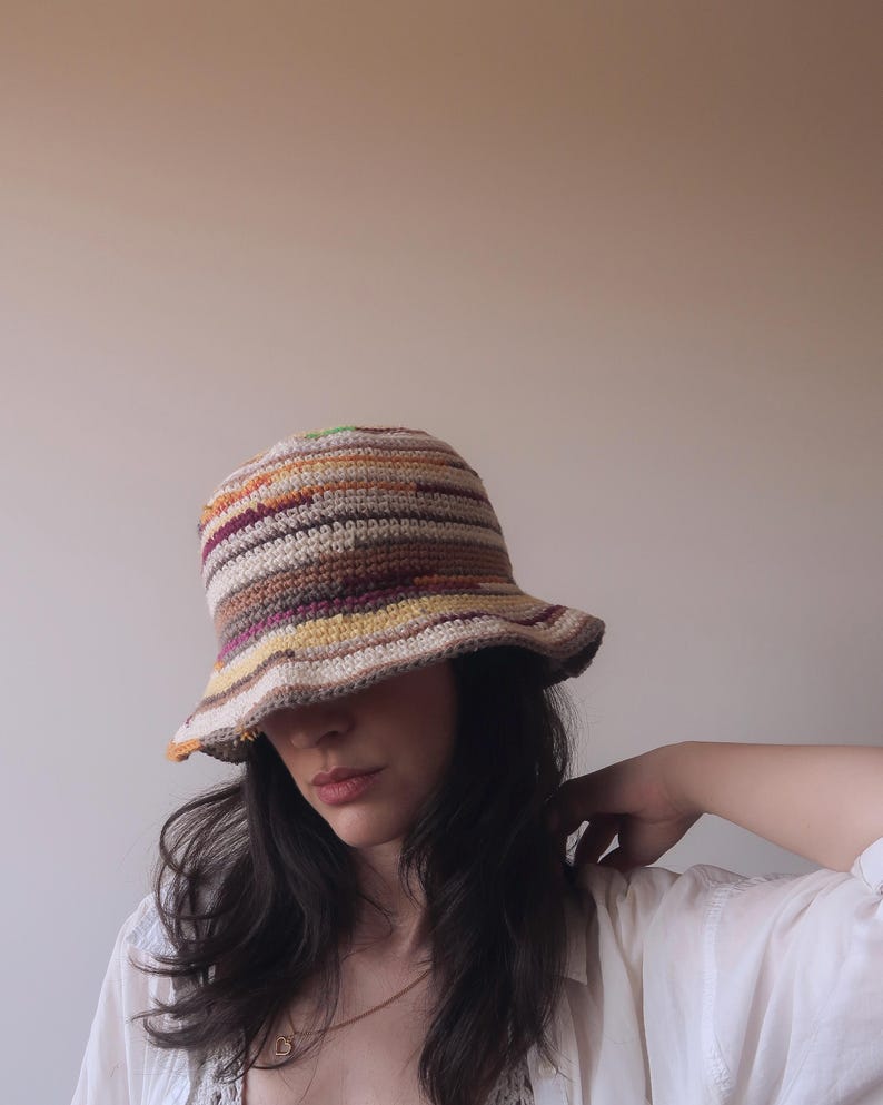 May include: A woman wearing a crocheted bucket hat with brown, orange, yellow, and white stripes. The hat is pulled down over her forehead, and she is looking down.