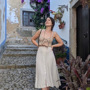 May include: A woman wearing a beige crochet top with a tie detail and a beige linen skirt with a button front. She is standing on a stone path in a cobblestone alleyway with a purple flowering vine growing on the wall.