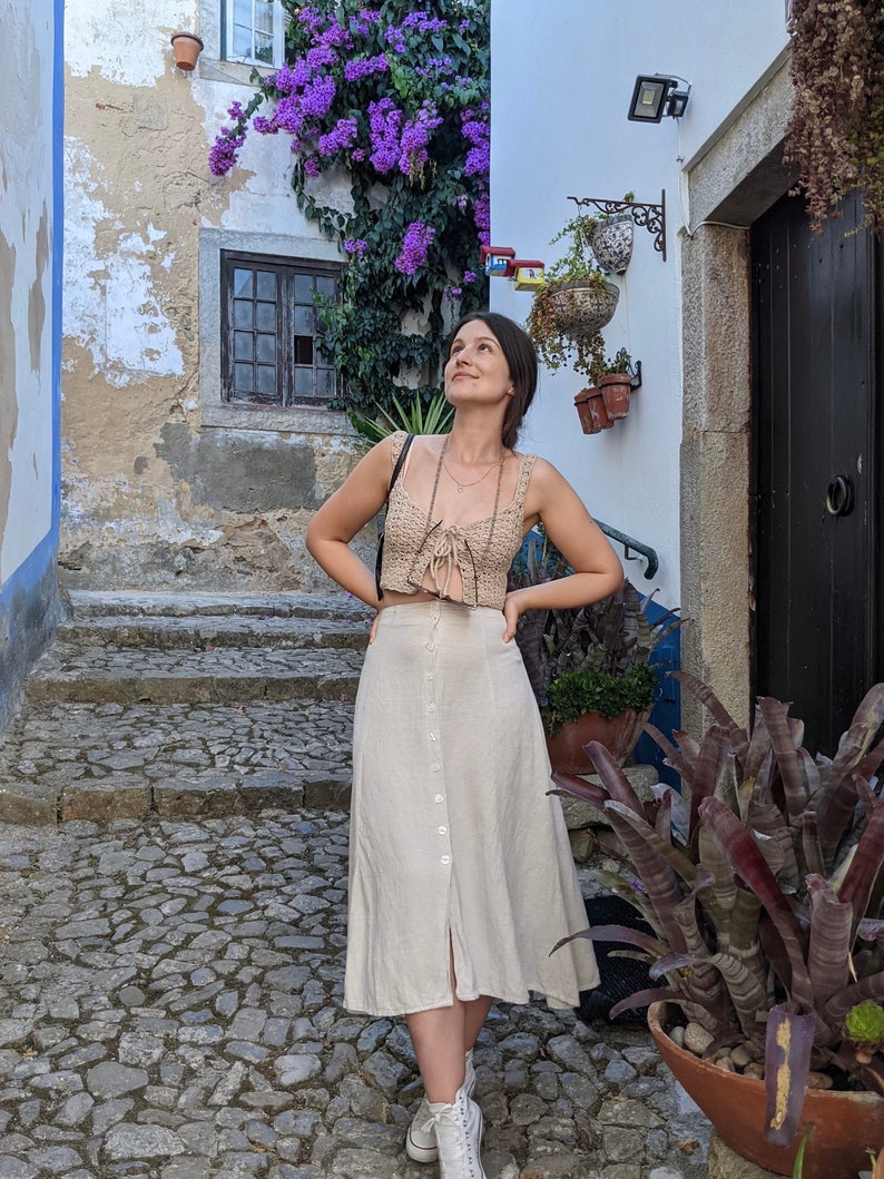 May include: A woman wearing a beige crochet top with a tie detail and a beige linen skirt with a button front. She is standing on a stone path in a cobblestone alleyway with a purple flowering vine growing on the wall.