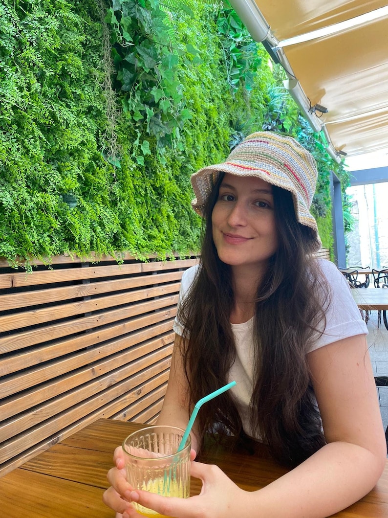 May include: A woman wearing a white shirt and a multi-colored crocheted bucket hat sits at a wooden table with a glass of orange juice and a blue straw.