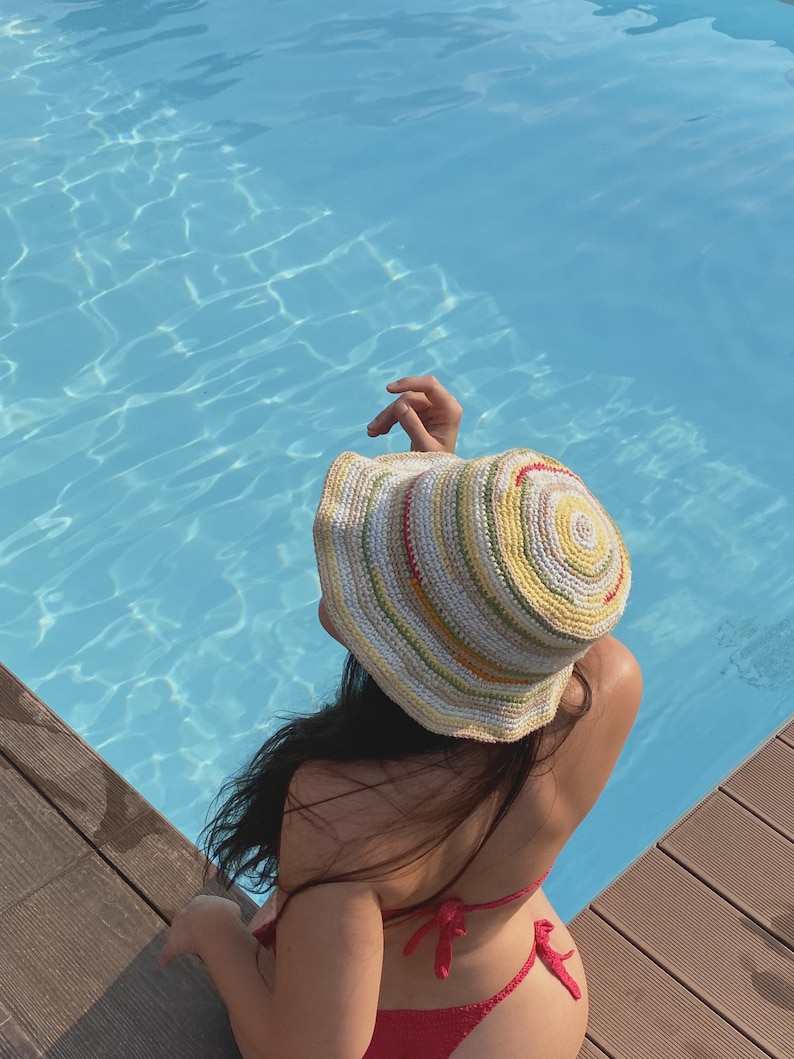 May include: A woman wearing a colorful crocheted bucket hat and a red crochet bikini top sits by a pool.
