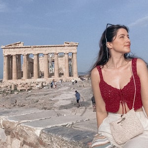 May include: A woman wearing a red crochet top and white pants sits on a stone wall in front of the Parthenon in Athens, Greece. She is wearing a straw bag and has a striped scarf draped over her shoulder.
