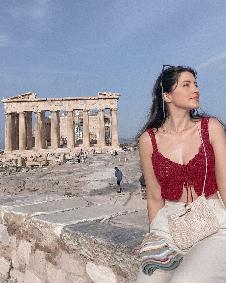 May include: A woman wearing a red crochet top and white pants sits on a stone wall in front of the Parthenon in Athens, Greece. She is wearing a straw bag and has a striped scarf draped over her shoulder.