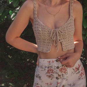 May include: A beige crochet crop top with a tie front and a heart-shaped pendant necklace. The top is paired with a white skirt with a floral print.