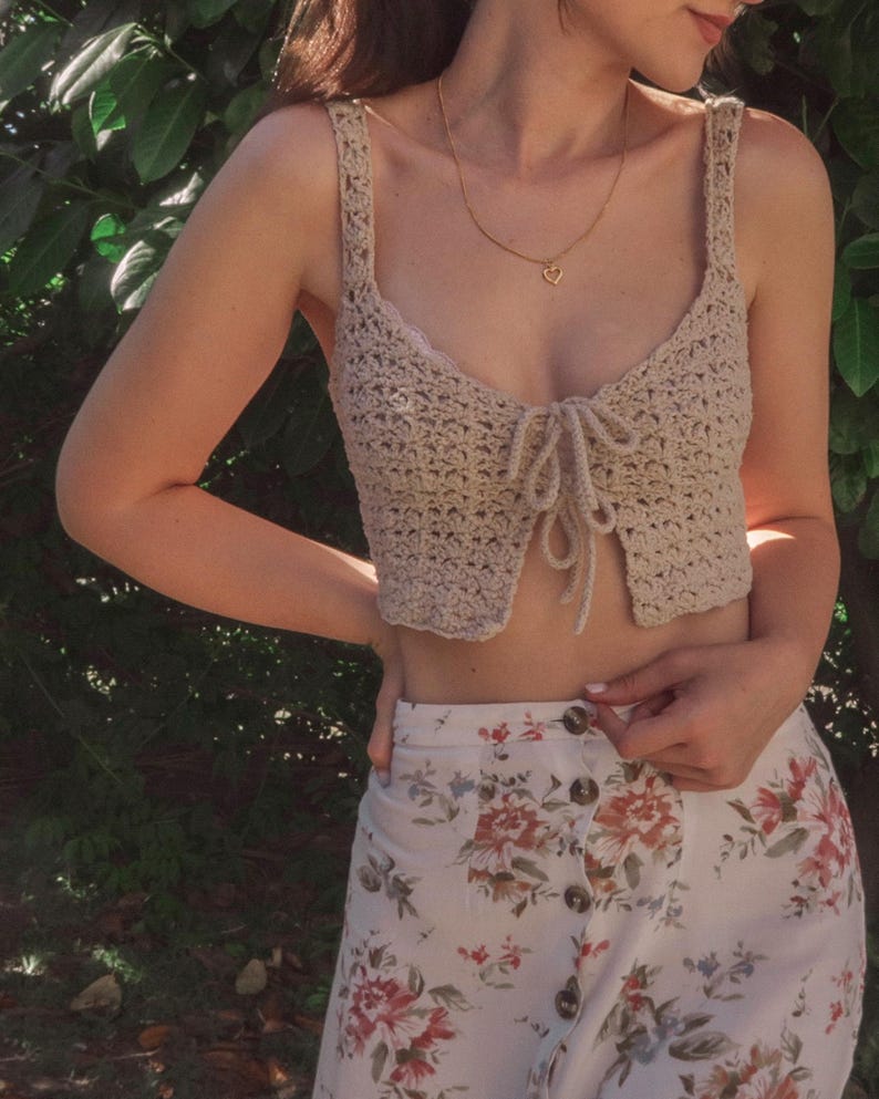 May include: A beige crochet crop top with a tie front and a heart-shaped pendant necklace. The top is paired with a white skirt with a floral print.