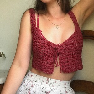 May include: A burgundy crochet crop top with a tie front detail. The top is made with a delicate stitch pattern and has a relaxed fit.
