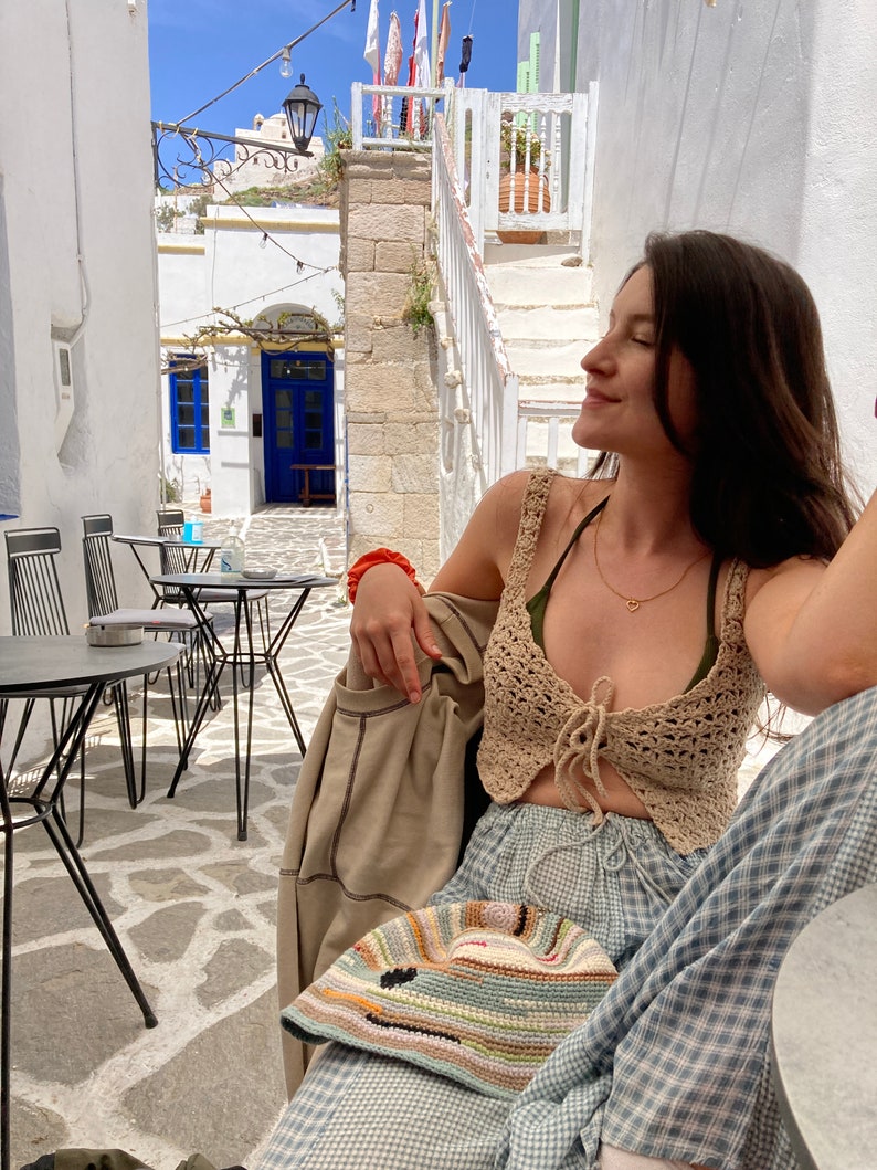 May include: A woman wearing a crocheted top with a tie front, blue and white plaid pants, and a colorful crocheted hat. She is sitting at a table outside in a white building with a blue door.
