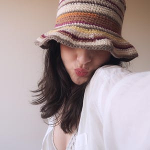 May include: A woman wearing a white shirt and a crocheted bucket hat with brown, orange, yellow, and purple stripes. The hat is pulled down low over her forehead, and she is looking at the camera with a playful expression.
