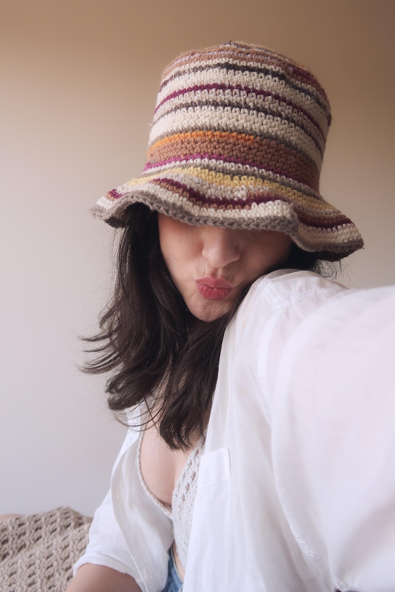 May include: A woman wearing a white shirt and a crocheted bucket hat with brown, orange, yellow, and purple stripes. The hat is pulled down low over her forehead, and she is looking at the camera with a playful expression.